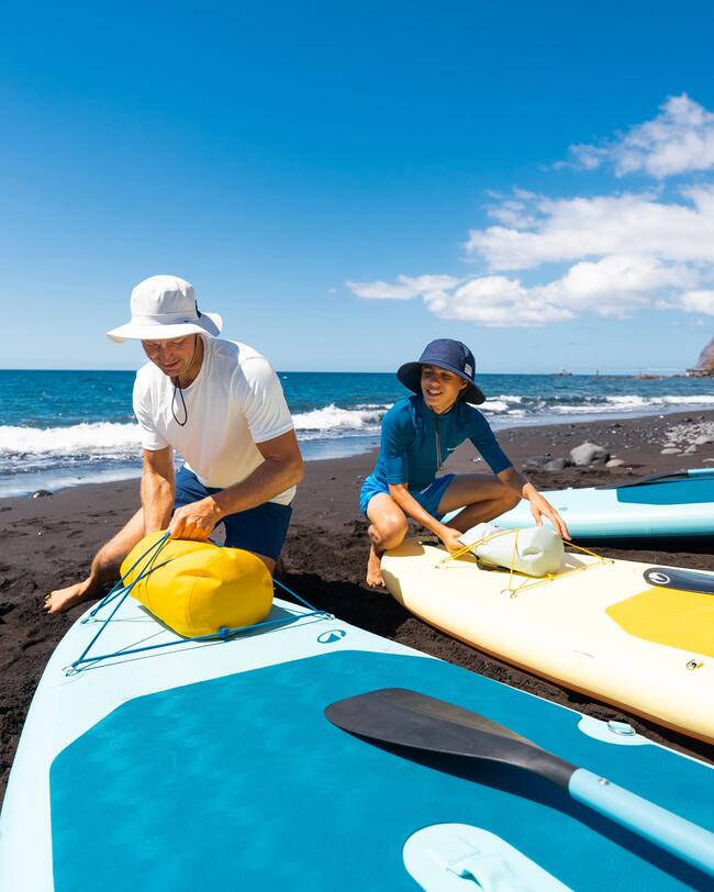 Zwei Personen in Hüten mit blauen und gelben Paddleboards am schwarzen Sandstrand unter blauem Himmel.