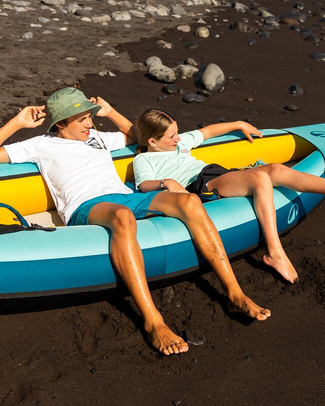 Zwei Personen entspannen sich in einem gelb-blauen Schlauchboot auf dunklem Sand mit Steinen.