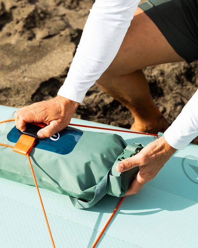 Hände befestigen grüne wasserdichte Tasche auf hellblauem Surfbrett; orangefarbene Seile; Sand im Hintergrund.
