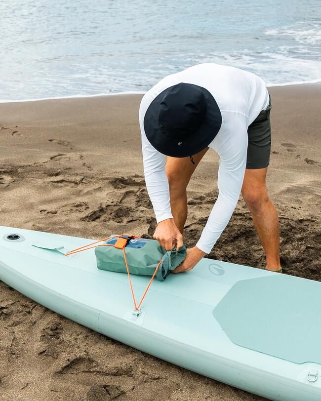 Person am Strand bindet Tasche auf mintfarbenem Paddleboard. Sandig, bewölkter Himmel, ruhige Wellen, entspannte Atmosphäre.