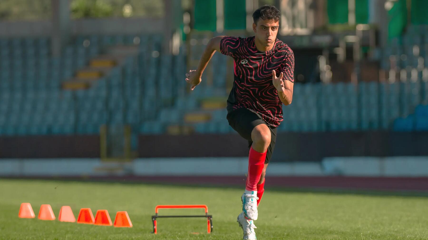 Ein junger Fußballspieler in einem schwarz-rot gemusterten Trainingsshirt absolviert auf einem Rasenplatz vor Stadionrängen ein Sprinttraining zwischen kleinen Hürden und Markierungshütchen.