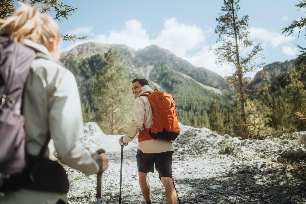 Muškarac sa narandžastim rancem i planinarskim štapovima hoda stazom kroz planinu, dok se u pozadini vidi žena koja ga prati.