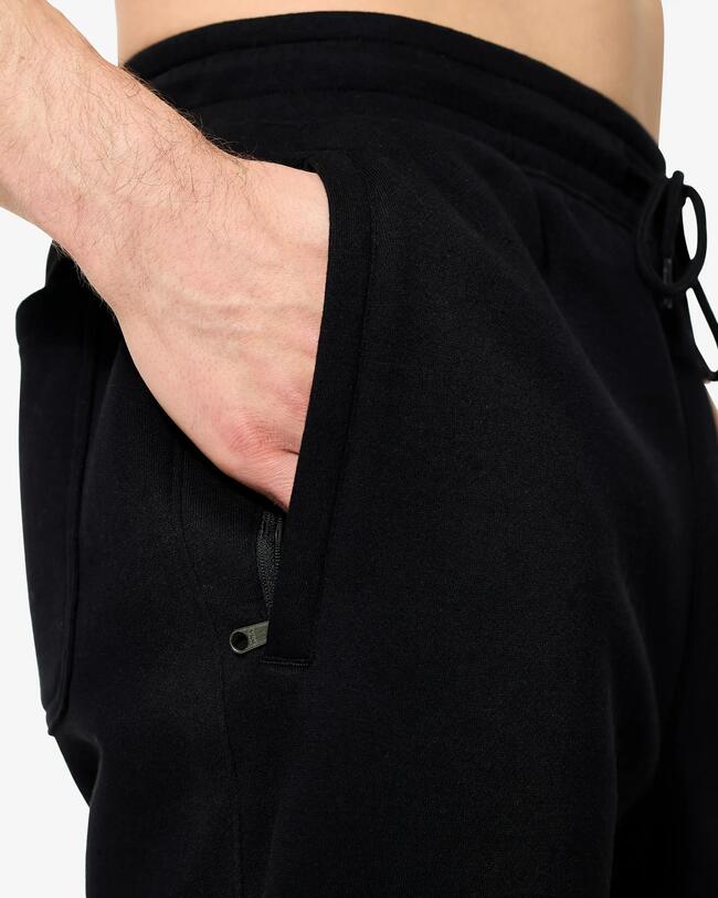 Nahaufnahme einer schwarzen Jogginghose mit Reißverschlusstasche und Kordelzug. Eine Hand steckt in der Tasche.
