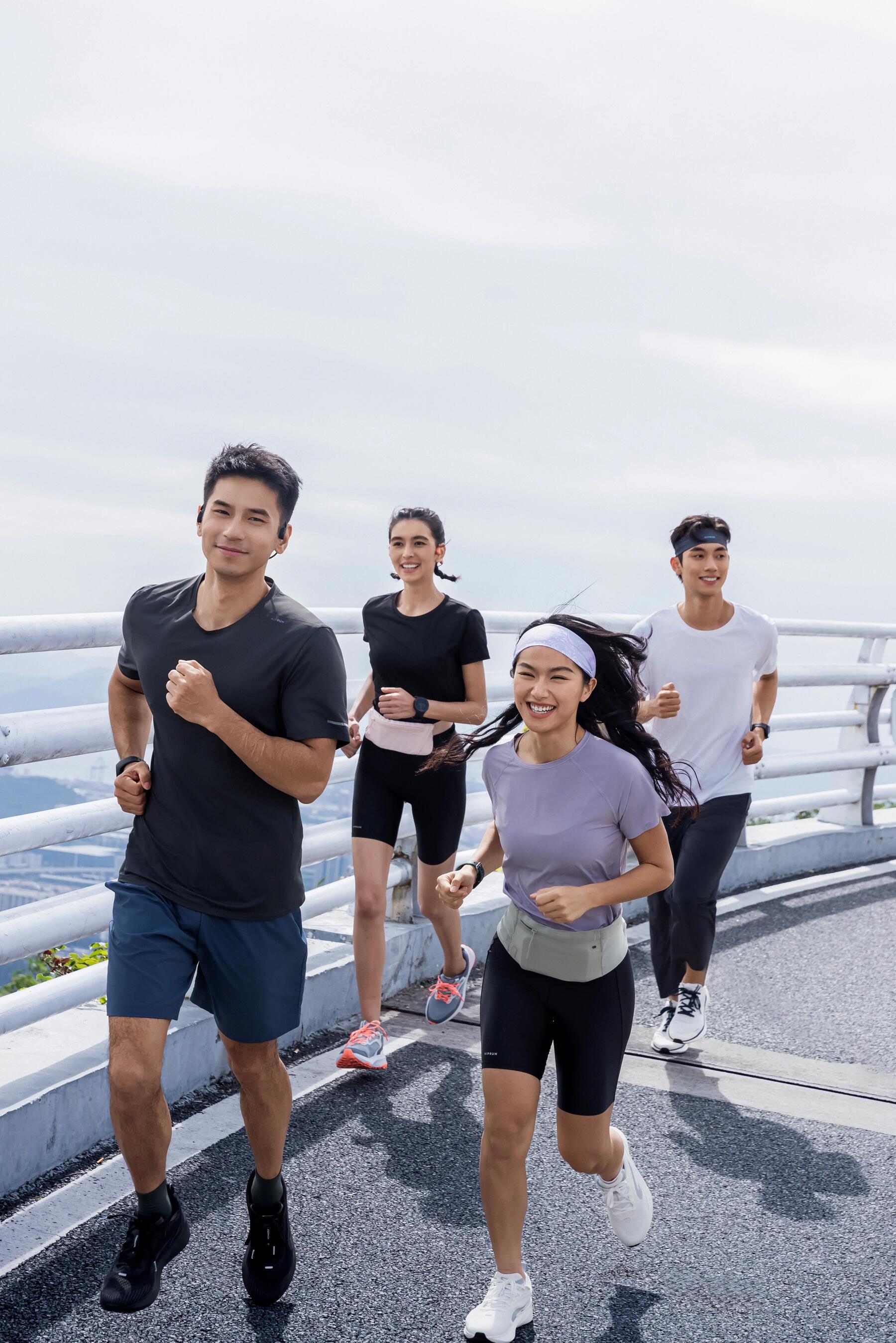 Vier junge Menschen joggen auf einer Asphaltstrecke, tragen sportliche Kleidung in Schwarz, Weiß und Violett, fröhliche Stimmung.