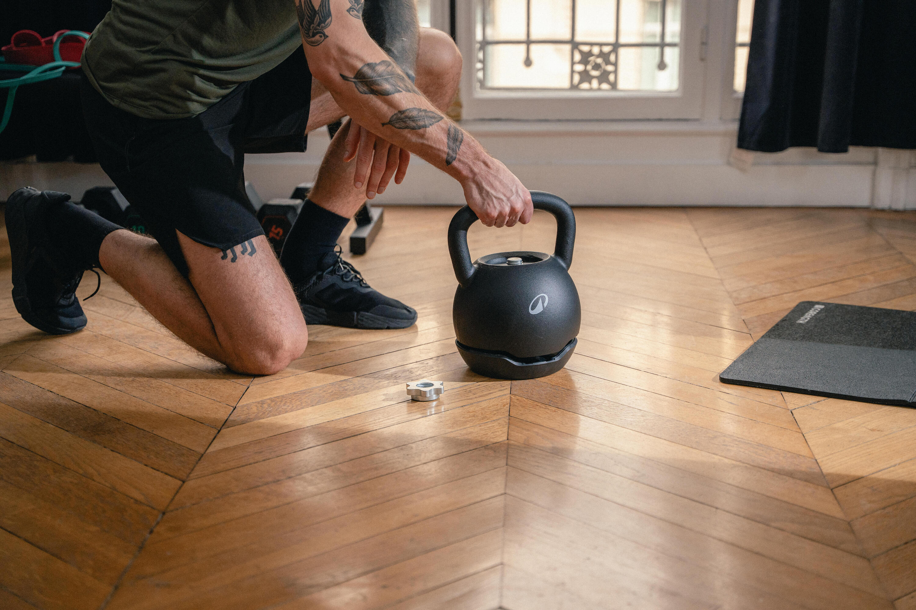 Kniender Mann mit Tätowierungen greift nach einer schwarzen Kettlebell auf einem Parkettboden.