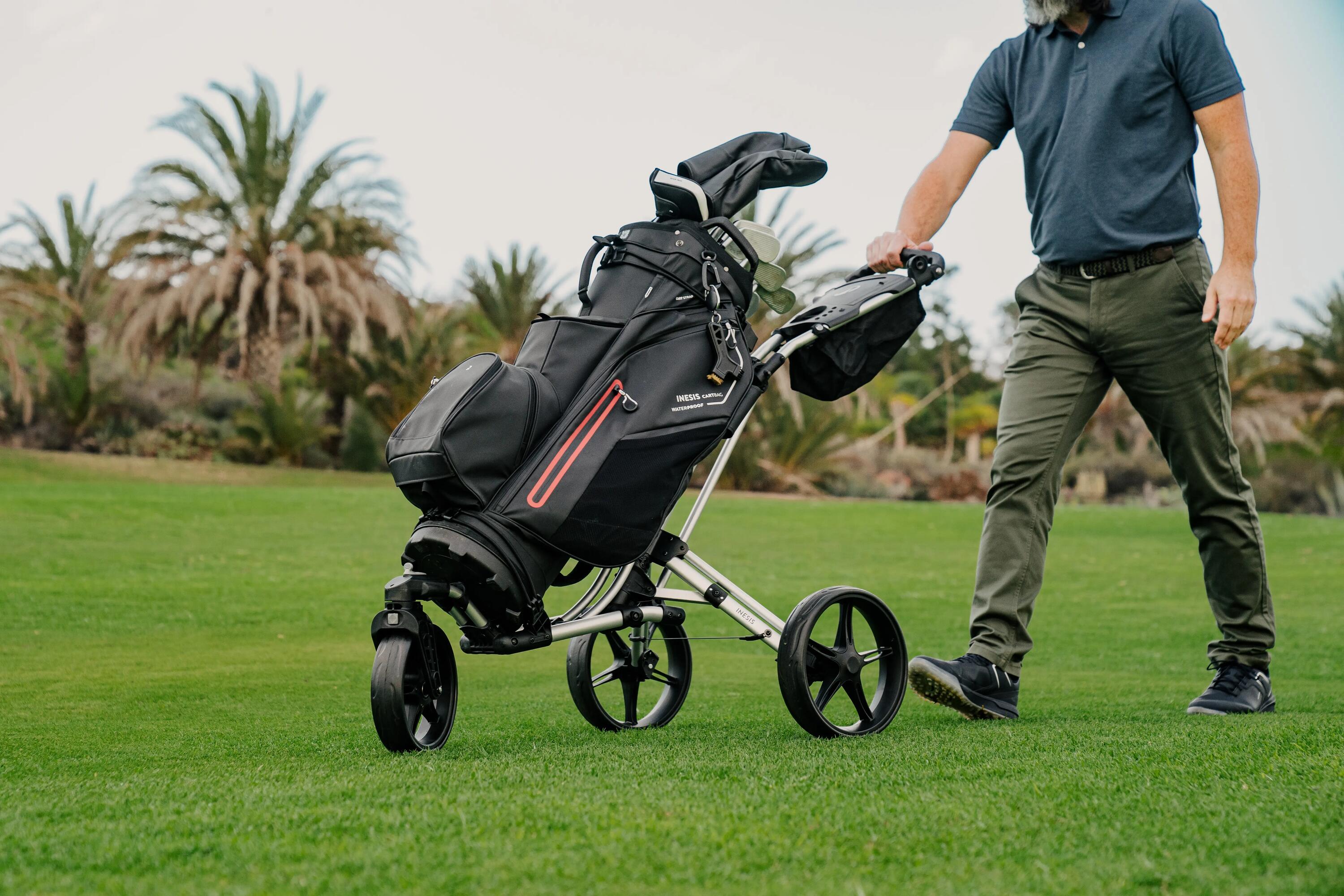 Ein bärtiger Mann, der ein kurzärmeliges blaues Poloshirt und olivgrüne Hosen trägt, schiebt einen dreirädrigen Golfwagen über einen üppig grünen Golfplatz. Seine linke Hand ruht auf dem Griff des silbernen Wagens, der eine große, gut ausgestattete schwarze Golftasche hält. In der Tasche sind mehrere Schläger, geschützt durch verschiedene Schlägerhauben, deutlich sichtbar. Der Mann, mit nach vorne ausgestrecktem linken Bein, scheint in Bewegung zu sein, möglicherweise zwischen den Löchern. Im Hintergrund sind mehrere Palmen vor einem sanften, hellen Himmel zu sehen, was auf einen tropischen oder subtropischen Ort für den Golfplatz hindeutet.
