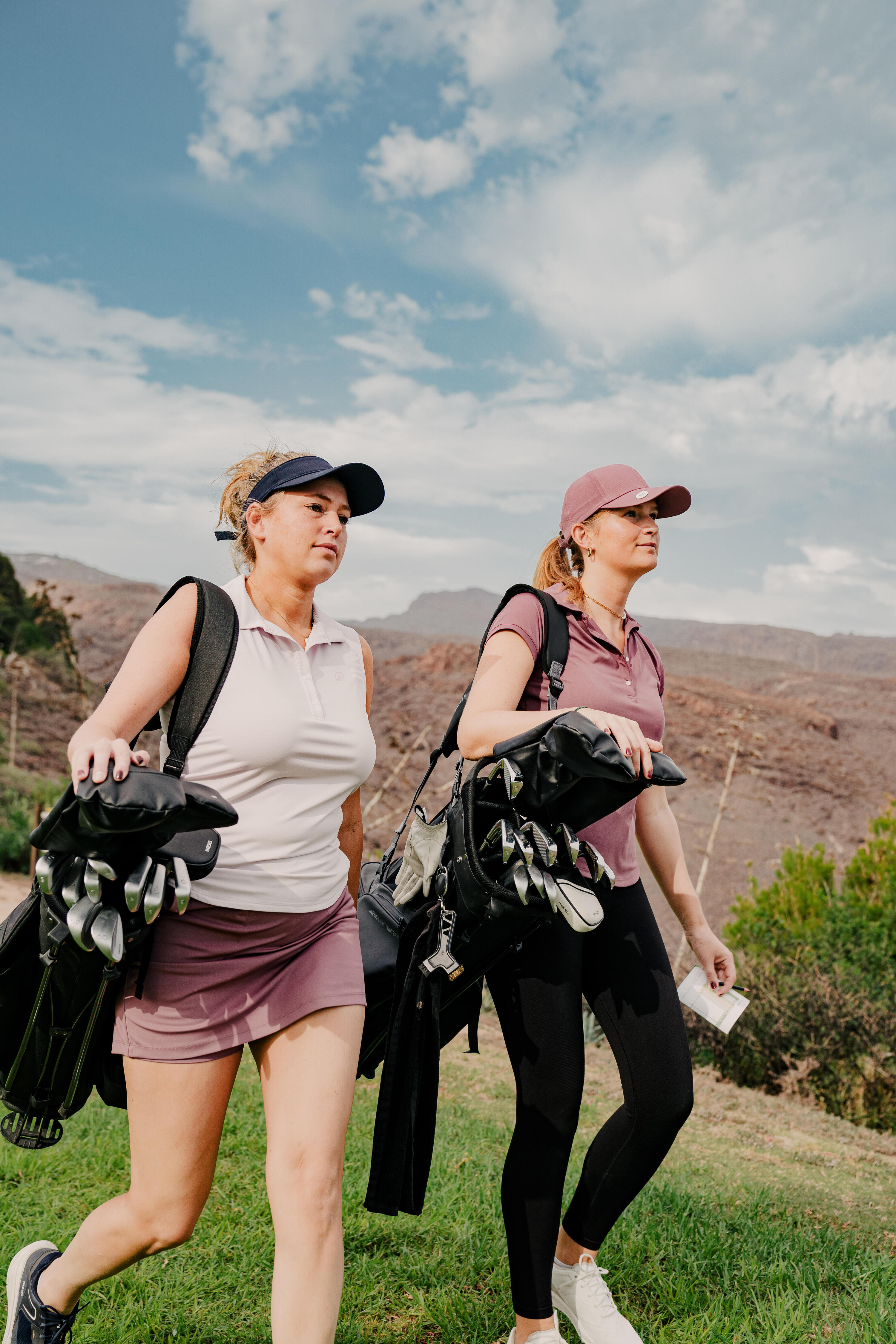 Zwei Frauen laufen im Freien mit zwei Golfbags über der Schulter über einen Golfplatz.