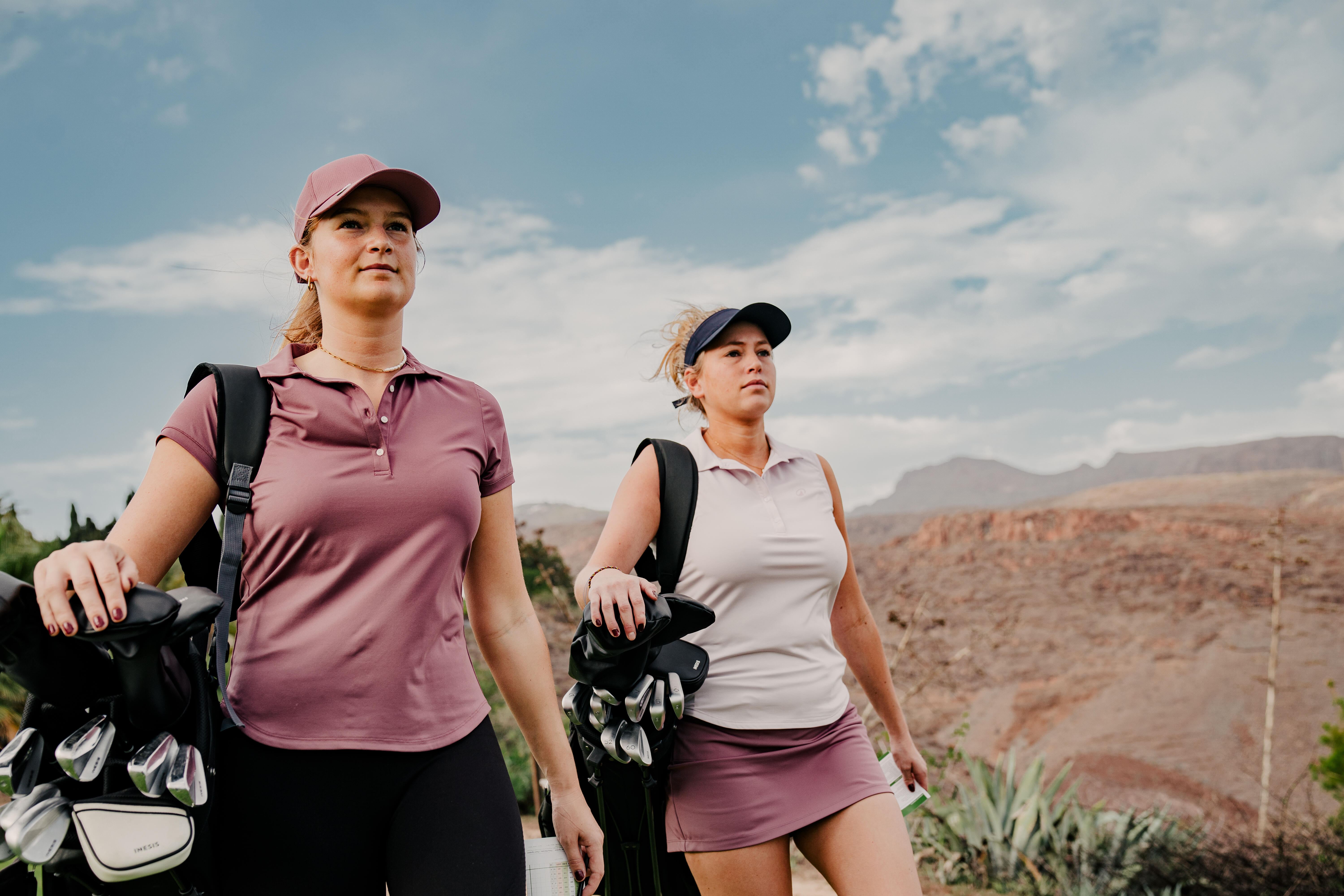 Two women in caps carrying golf bags across a rocky course