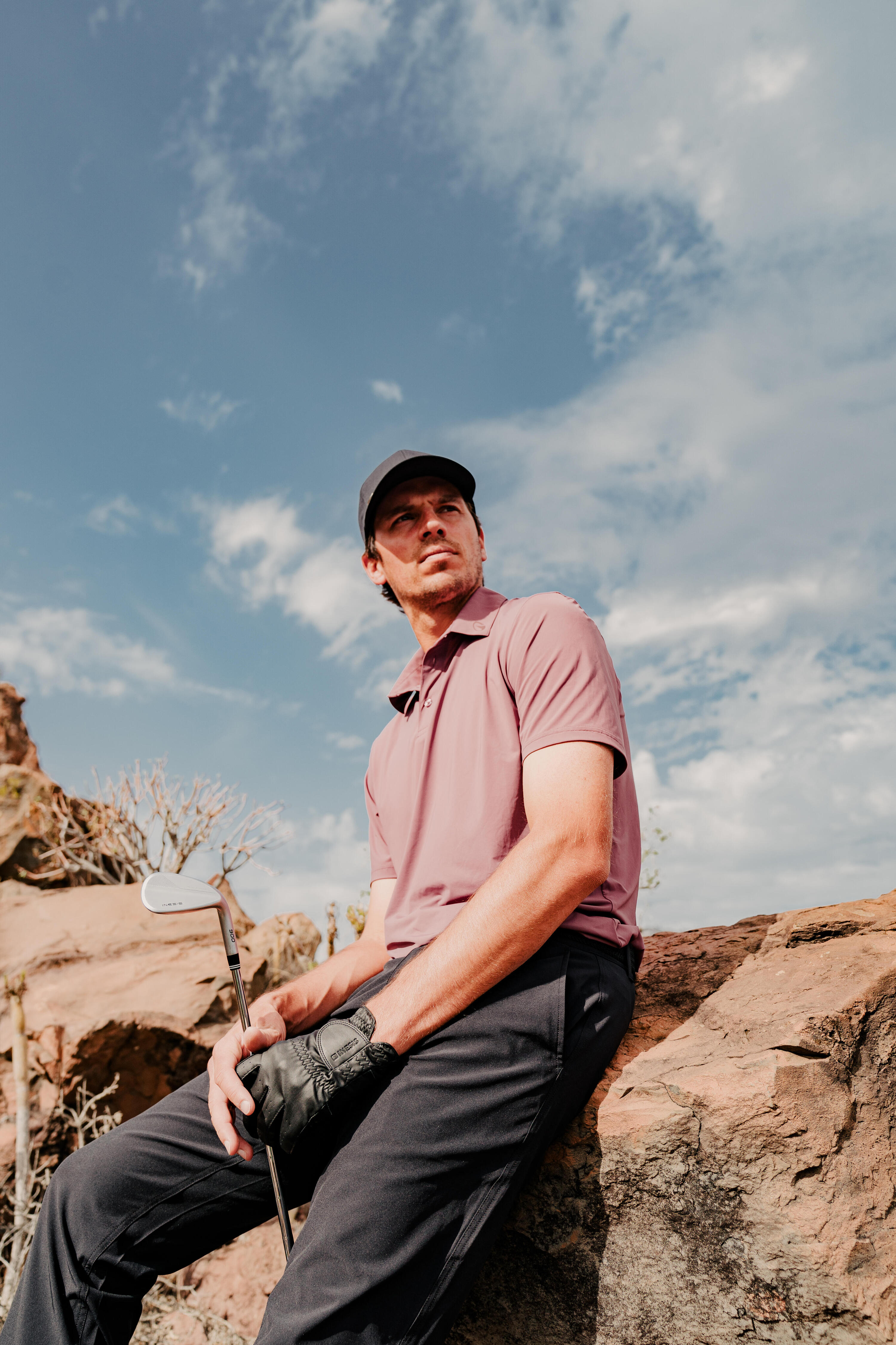 Ein Golfer in weinrotem Poloshirt, schwarzer Hose und dunkler Cap sitzt entspannt auf einem Felsen in einer rauen Landschaft, blickt in die Ferne und hält dabei locker sein Golf-Eisen in der Hand.