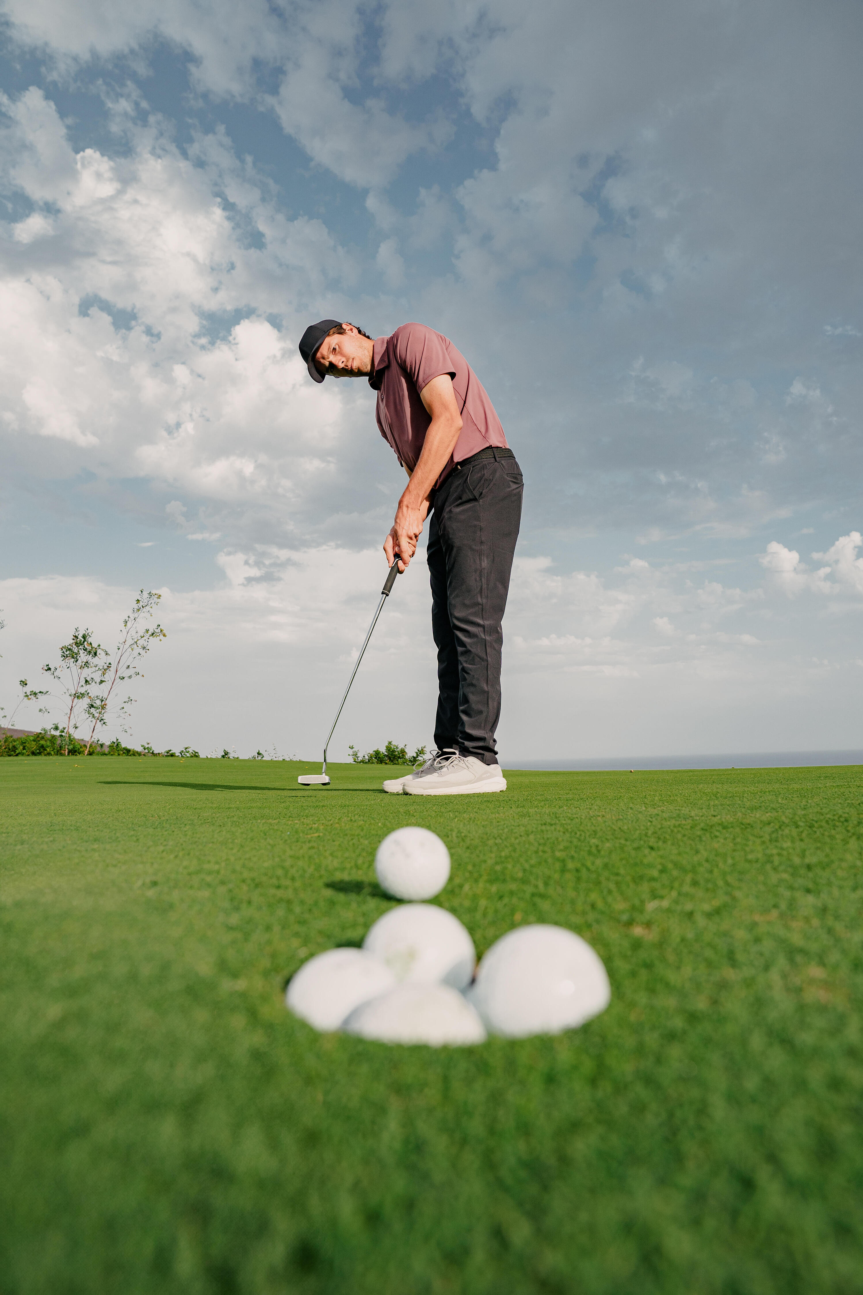 Ein Golfer in weinrotem Poloshirt und schwarzer Hose steht auf einem perfekt gepflegten Grün unter weitem Himmel und bereitet sich auf einen Putt vor, während im direkten Vordergrund mehrere Golfbälle bereits im Loch liegen.