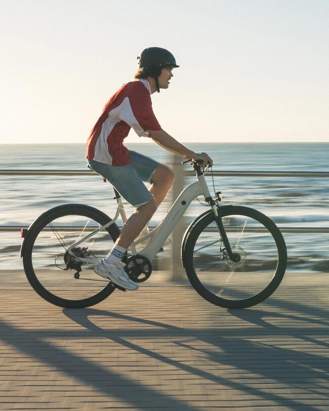 Mann mit schwarzem Helm und rotem T-Shirt fährt auf weißem E-Bike eine Uferpromenade entlang.