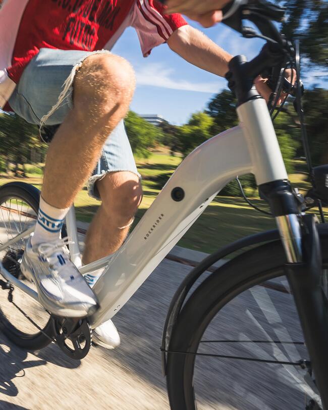 Person in Jeans und roten Hemd auf weißem E-Bike, fährt auf sonnigem Weg, Schatten und Bäume im Hintergrund.