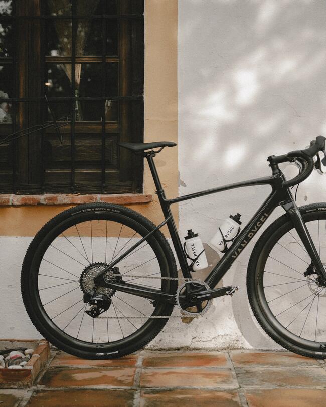 Schwarzes Fahrrad lehnt an einer cremeweißen Mauer auf rotbraunen Fliesen, mit Schatten von Bäumen auf der Wand.
