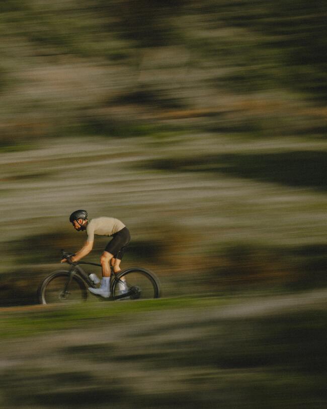 Radfahrer in verschwommener grüner Landschaft, trägt Helm und dunkle Kleidung, bewegt sich schnell seitlich über Gras.