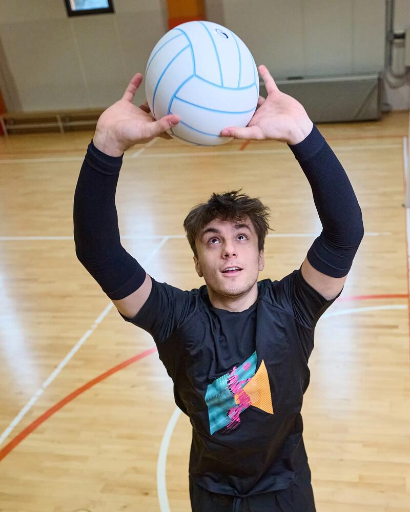 Mann mit schwarzen Klamotten prtischt einen Volleyball in einer Halle.