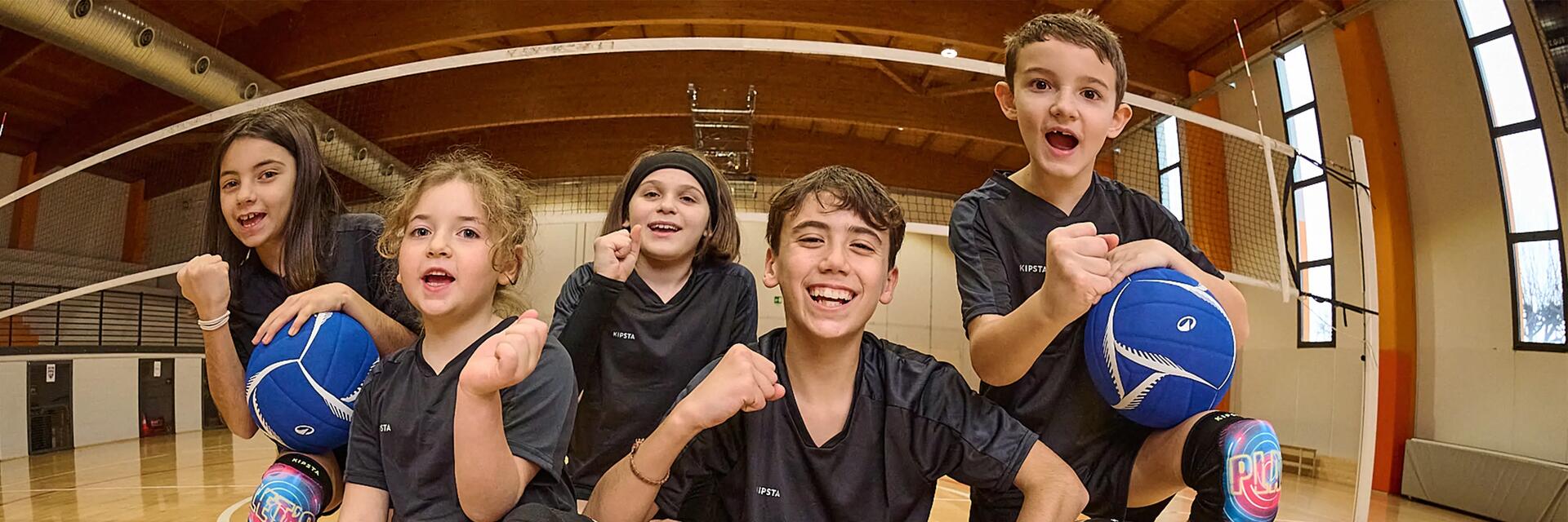 Auf diesem Bild sieht man fünf fröhliche Kinder in dunkler Sportkleidung, die mit blauen Bällen auf dem Holzboden einer Sporthalle sitzen und in die Kamera jubeln.