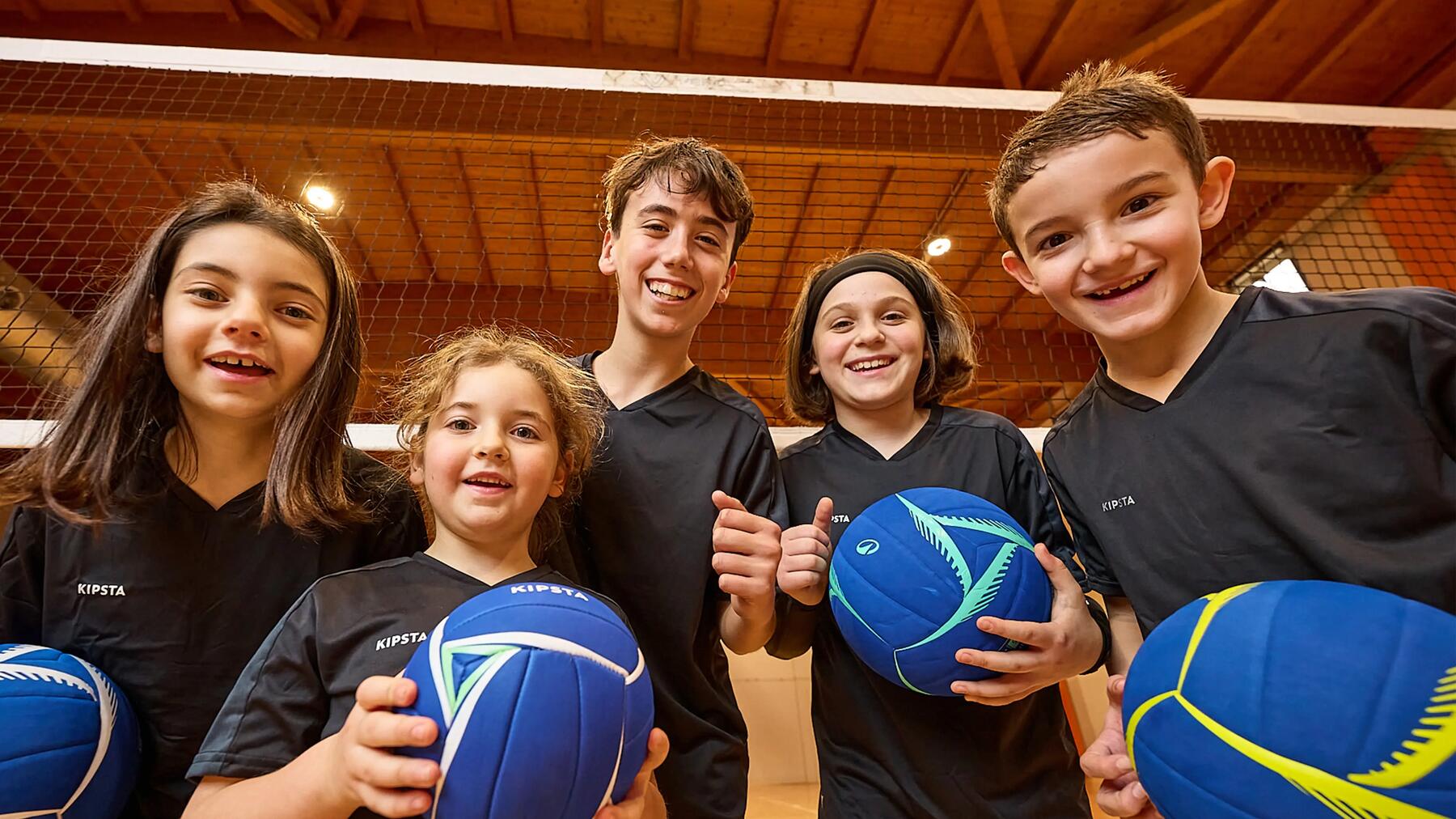 Fünf Kinder haben jeweils einen Volleyball in der Hand und lächeln in die Kamera. Sie befinden sich in einer Sprothalle vor einem Volleyballnetz.