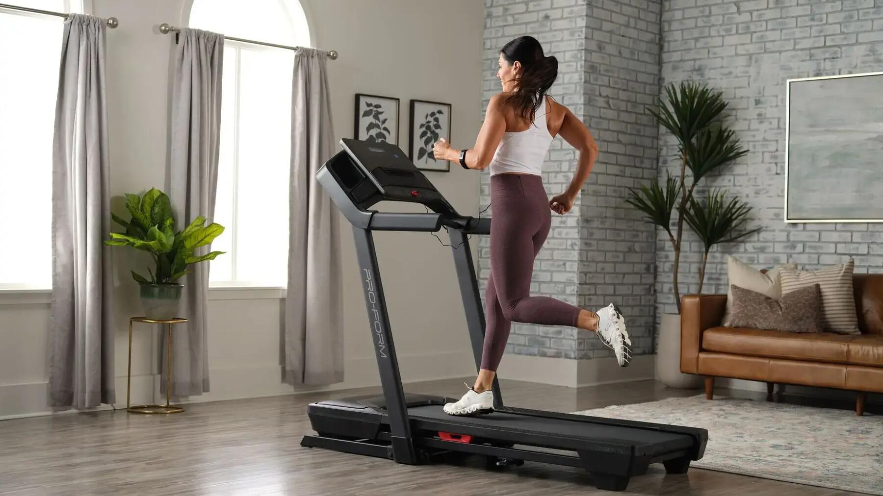 Frau mit weißem Top, lilanen Leggings und weißen Schuhen auf schwarzem Laufband im Wohnzimmer.