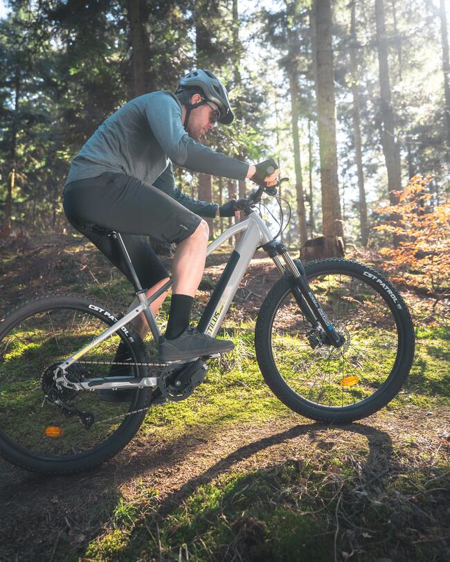 Mann in dunklem Sportoutfit mit Helm und Sonnenbrille fährt mit grauem Mountainbike durch sonnigen Wald
