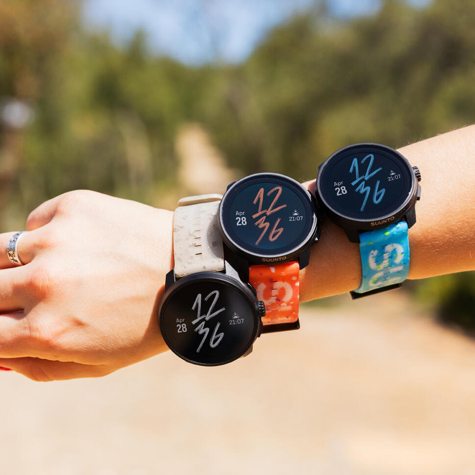 Tri športne ure Suunto na zapestju osebe, s paščki v krem, oranžni in modri barvi.