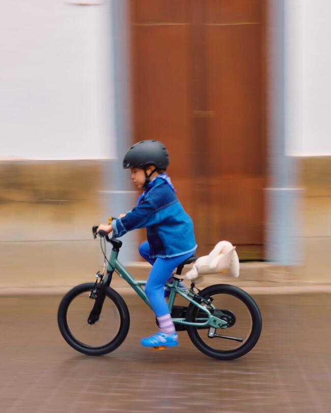 Ein Kind mit schwarzem Helm fährt auf grünem Fahrrad schnell eine Straße entlang. Im Hintergrund Hausfassade unscharf