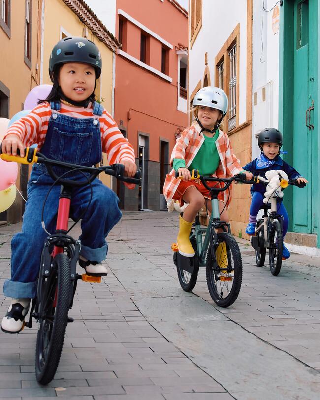 Drei Kinder fahren verstzt auf bunten Fahrrädern eine Straße mit bunten Fassaden lang