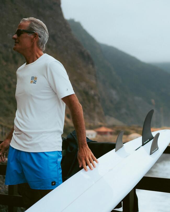 Mann in blauem Shorts und weißem Shirt mit Surfbrett, Berge im Hintergrund, bewölkter Himmel.