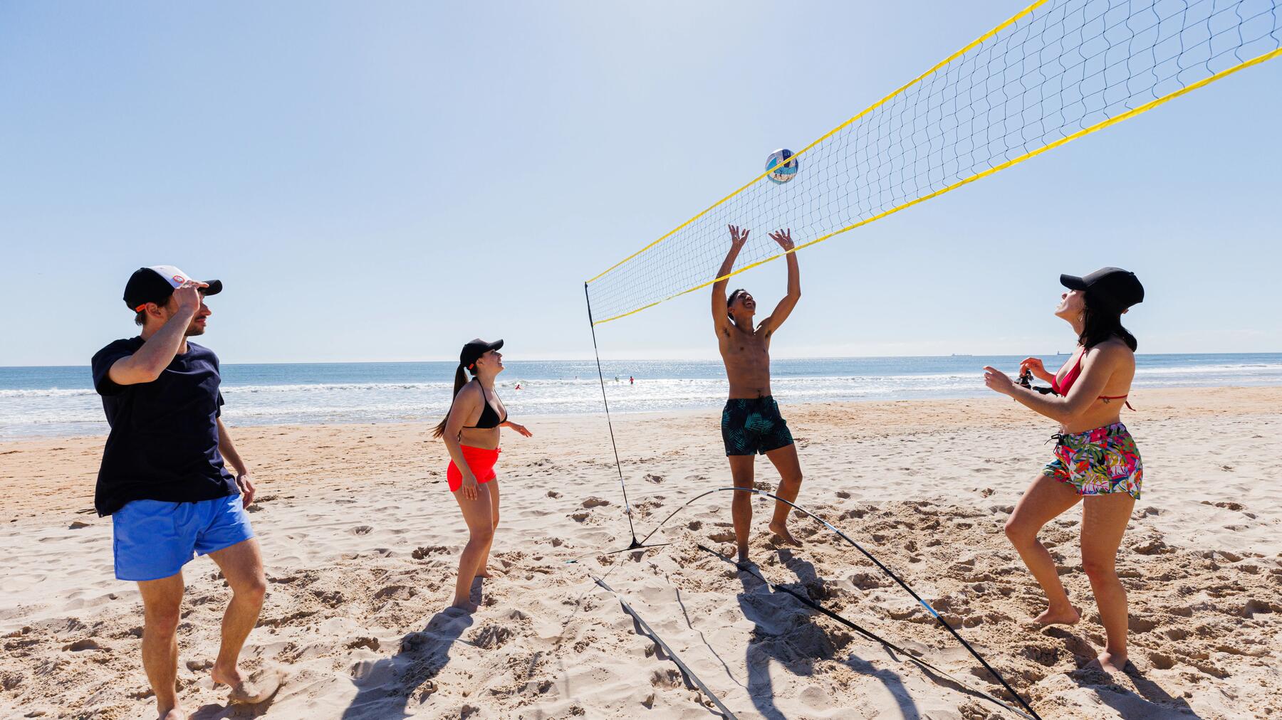 Eine Gruppe von zwei Männern und zwei Frauen spielt bei strahlendem Sonnenschein am Strand Beachvolleyball über ein Netz direkt am Meer.