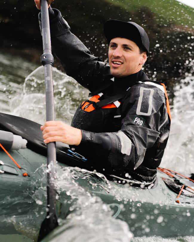 Person in schwarzer Ausrüstung paddelt im grauen Kajak; spritzendes Wasser, grüne Landschaft im Hintergrund.