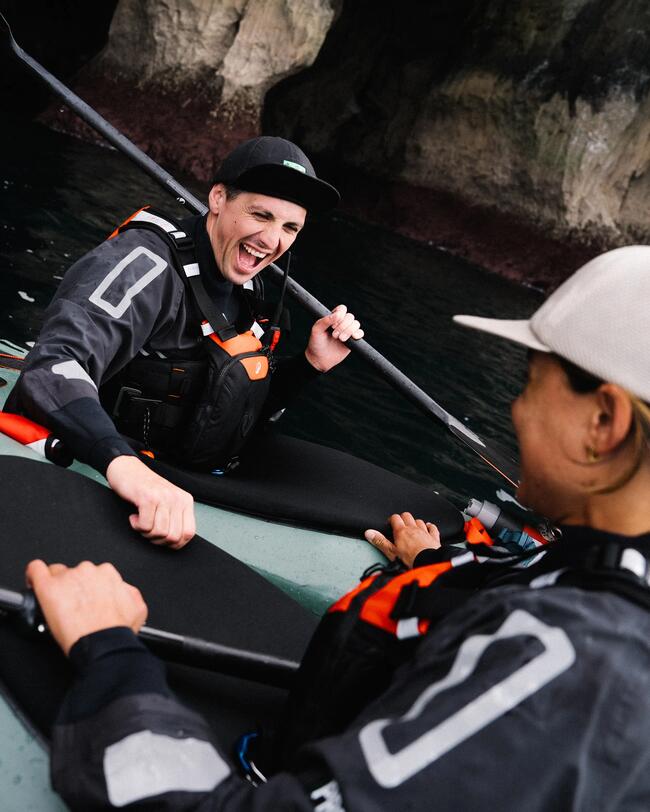 Zwei lachende Personen in schwarzen Jacken und Kappen auf einem Boot auf dunklem Wasser vor Felsen.