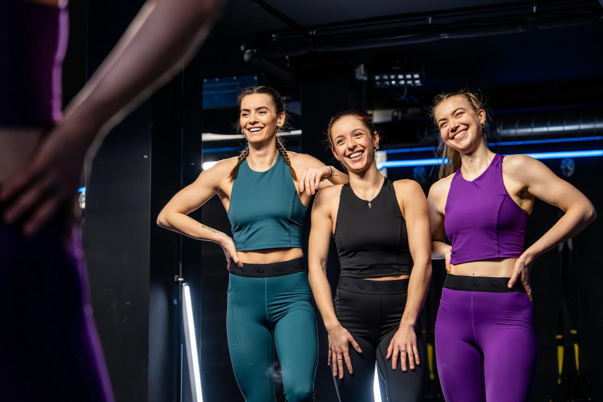 3 hölgy különböző Domyos márkájú sport felsőben és fitnesz nadrágban mosolyogva áll a fitneszteremben.