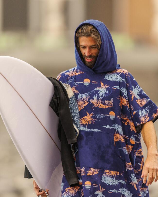  Ein lächelnder Mann mit Bart und nassen Haaren trägt einen blauen, gemusterten Poncho mit Kapuze und hält ein Surfbrett, was auf schnelles und "Natürlich trocken" nach dem Wassersport hinweist.