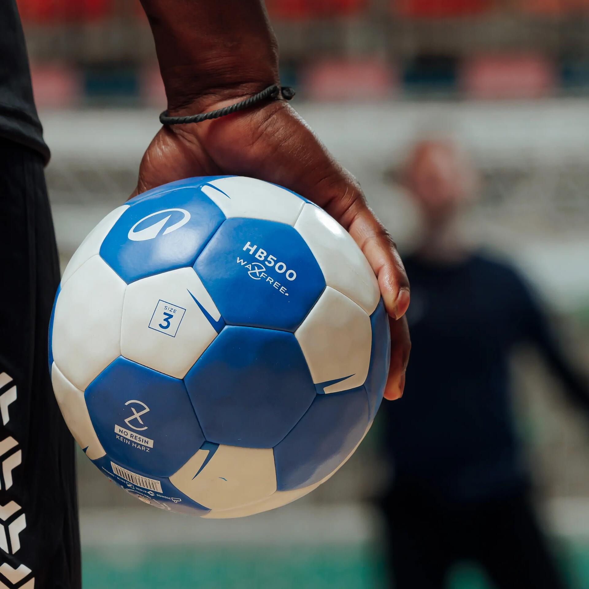 Naheaufnahme Hand mit blau-weißem Handball im Spielfeld, Spieler im Hintergrund, sportliche Atmosphäre.