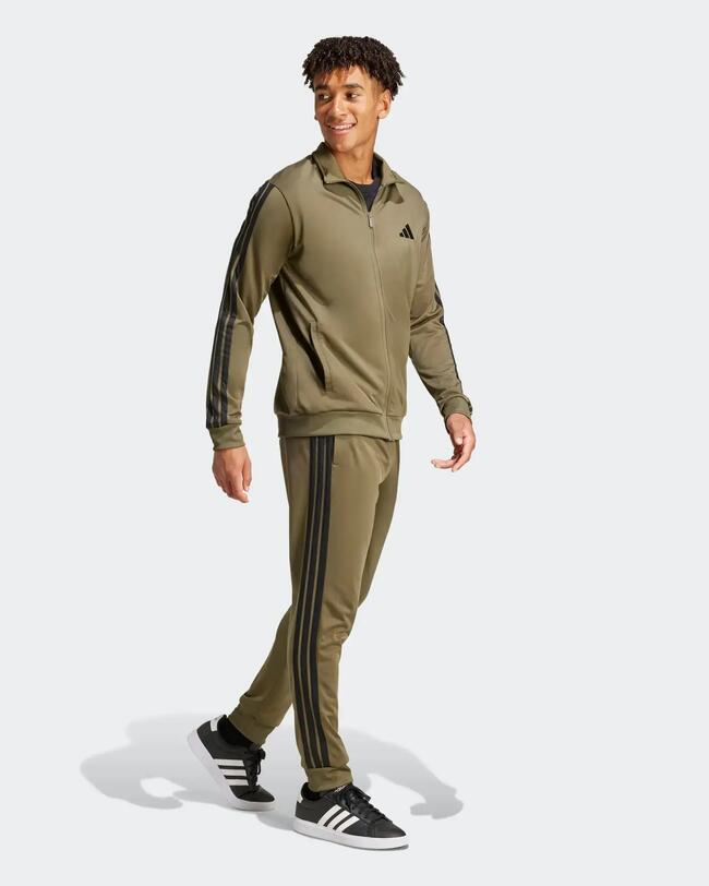 Lächelnder Mann in khakifarbenem Adidas-Trainingsanzug mit schwarzen 3-Streifen, beim Gehen frontal.