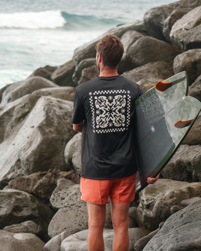 Person in schwarzem Shirt und orangefarbenen Shorts, hält Surfbrett, vor grauen Felsen, Meer im Hintergrund.