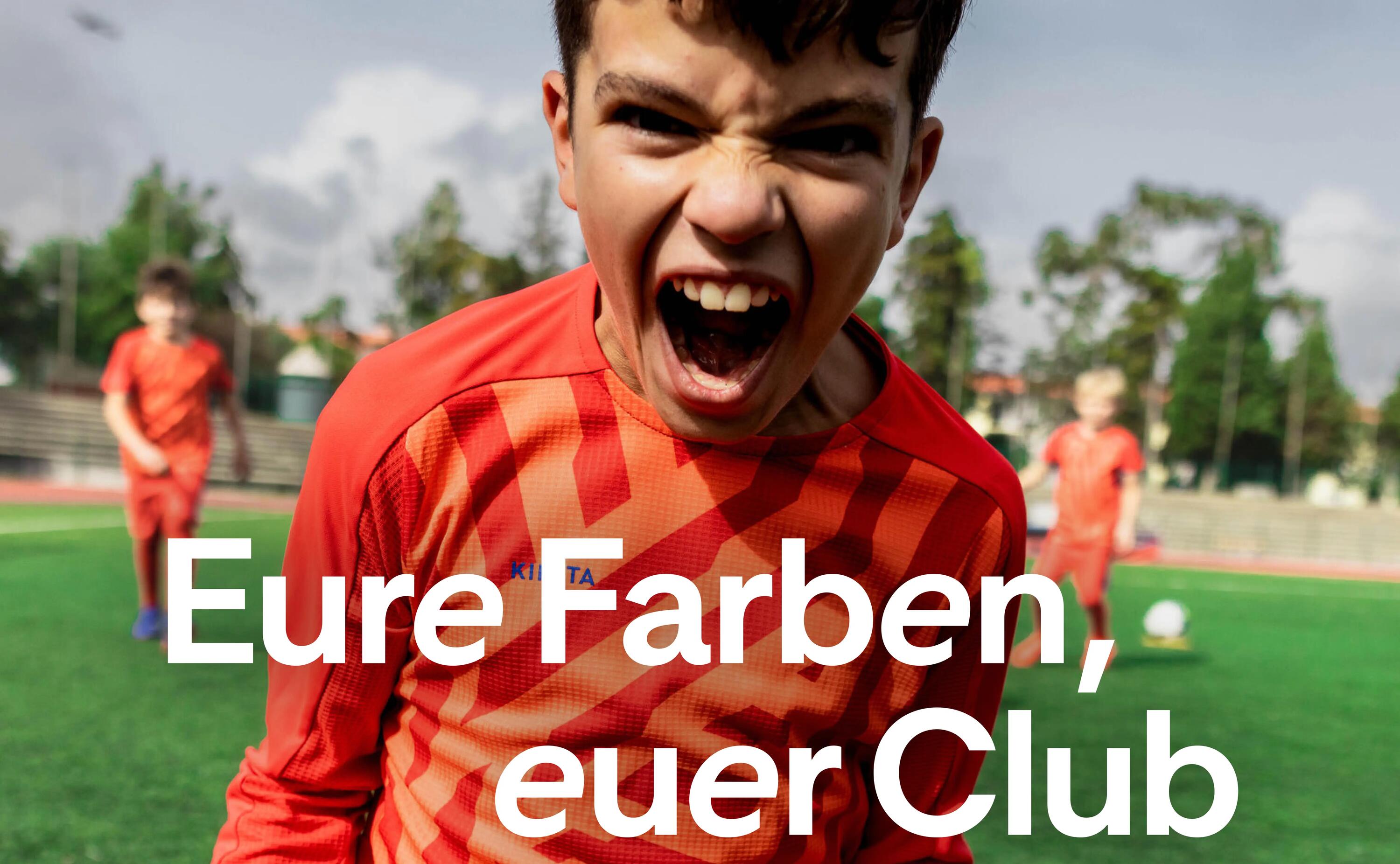 Junge im roten Fußballtrikot schreit begeistert, umgeben von Teamkameraden auf grünem Spielfeld.