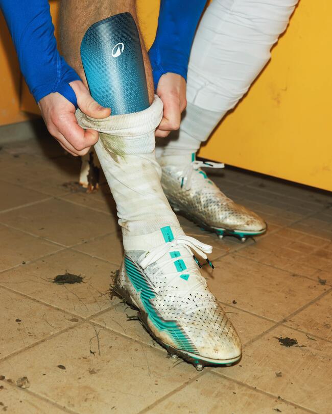 Weiß-blaue Fußballschuhe und Schienbeinschoner, Matschflecken auf Fliesenboden, gelber Hintergrund.