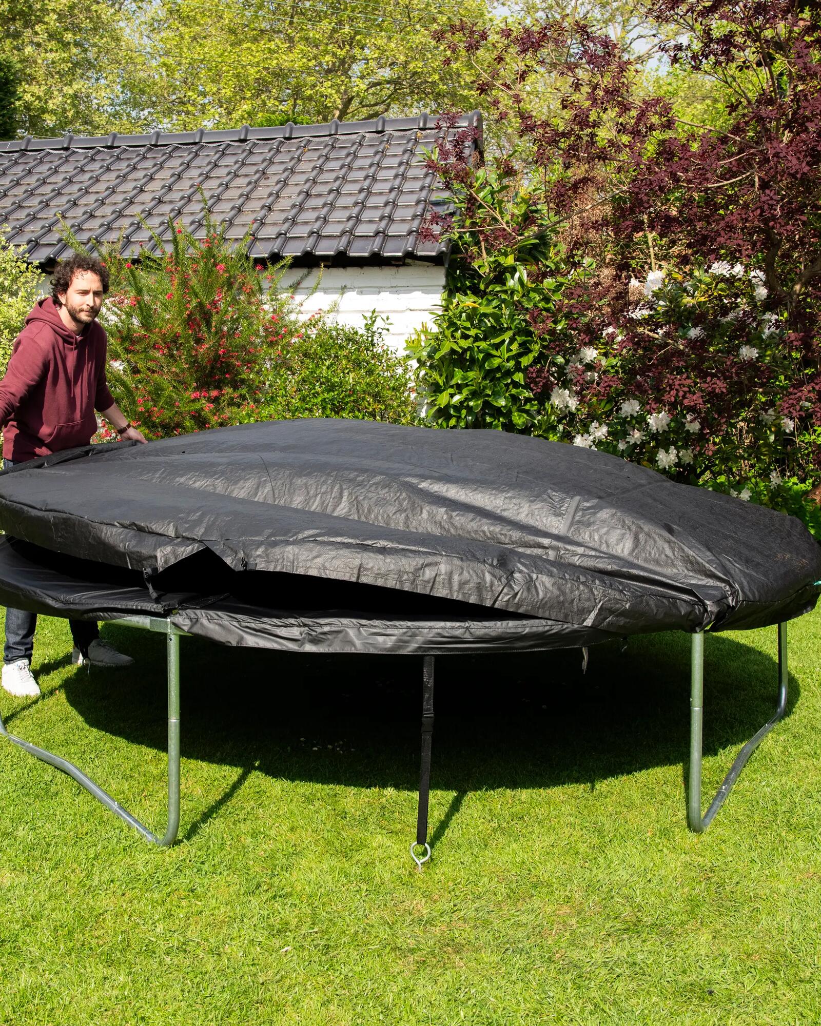 Ein Mann breitet eine schwarze Abdeckplane über ein großes Trampolin auf einer grünen Rasenfläche im Garten aus.