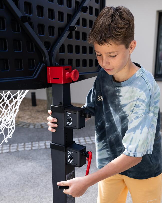 Junge in Batik-Shirt stellt schwarzen Basketballkorb ein, rotes Einstellelement, grauer Boden, weißes Netz.
