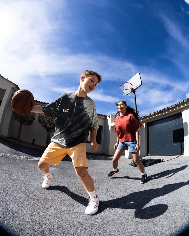 Junge und Mädchen spielen Basketball auf Einfahrt, blauer Himmel, gelbe Shorts, rotes Oberteil, Sonnenschein, Basketballkorb.
