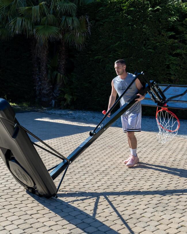 Mann schiebt schwarzen Basketballkorb auf gepflastertem Platz; Palmen und Hecken im Hintergrund.