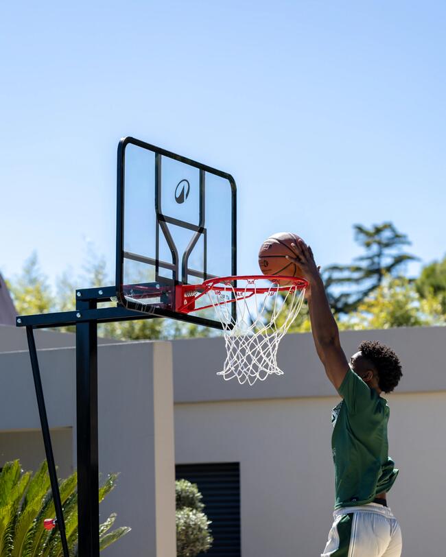 Basketballspieler im grünen Trikot macht Dunk. Blauer Himmel, grüne Bäume, weißer Korb.