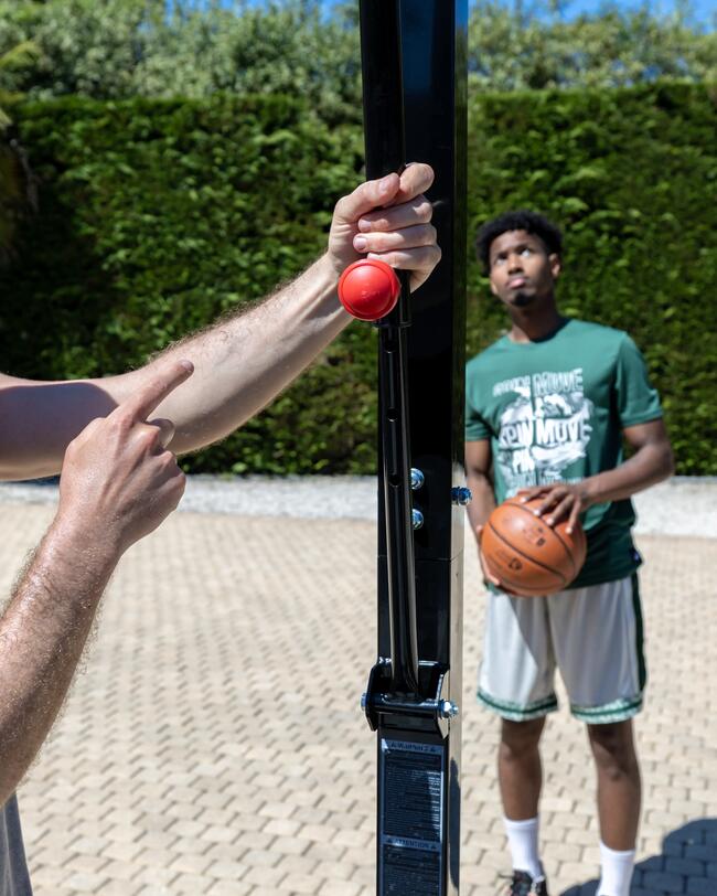 Hand zeigt auf roten Knopf an schwarzem Pfosten, Basketballspieler im Hintergrund, grüner Himmel, grauer Boden.