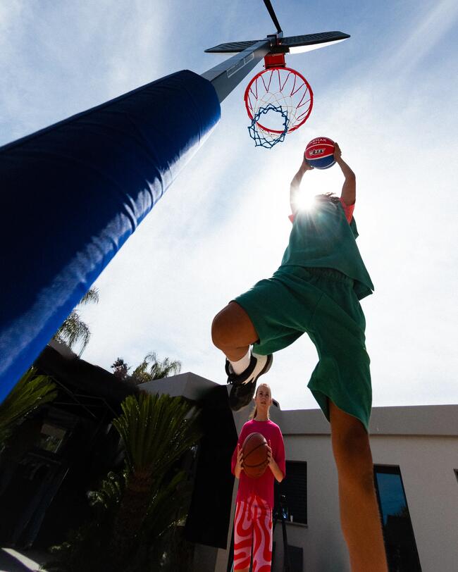 Basketballkorb-Sicht aus Froschperspektive, Spieler in Grün springt, blauer Himmel, helle Sonne, Palme im Hintergrund.