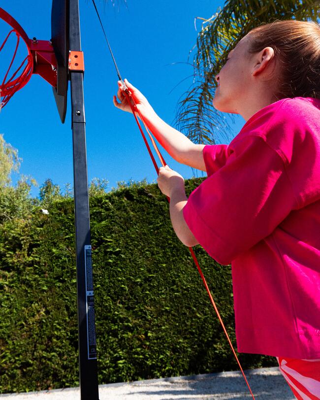 Mädchen in pinkem Shirt spannt rote Schnur am Basketballkorb, Hintergrund mit blauem Himmel und grüner Hecke.