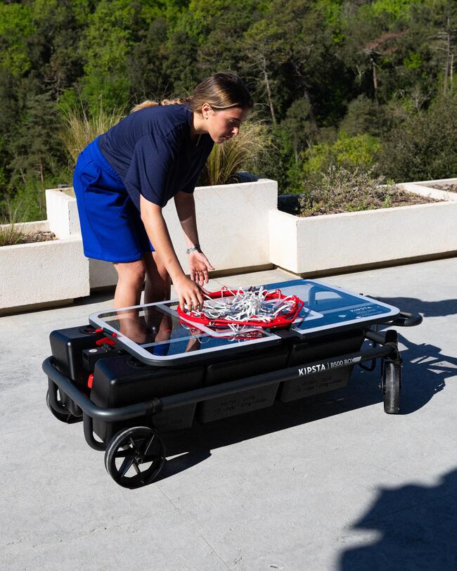 Mädchen befestigt rot-weißen Basketballkorbmontagesatz auf schwarzem Rollwagen. Blauer Rock, sonniger Tag, Umbegung mit Grünpflanzen.