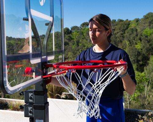 Mädchen justiert Basketballkorb, roter Ring, weißes Netz, blauer Himmel, grüne Landschaft im Hintergrund.