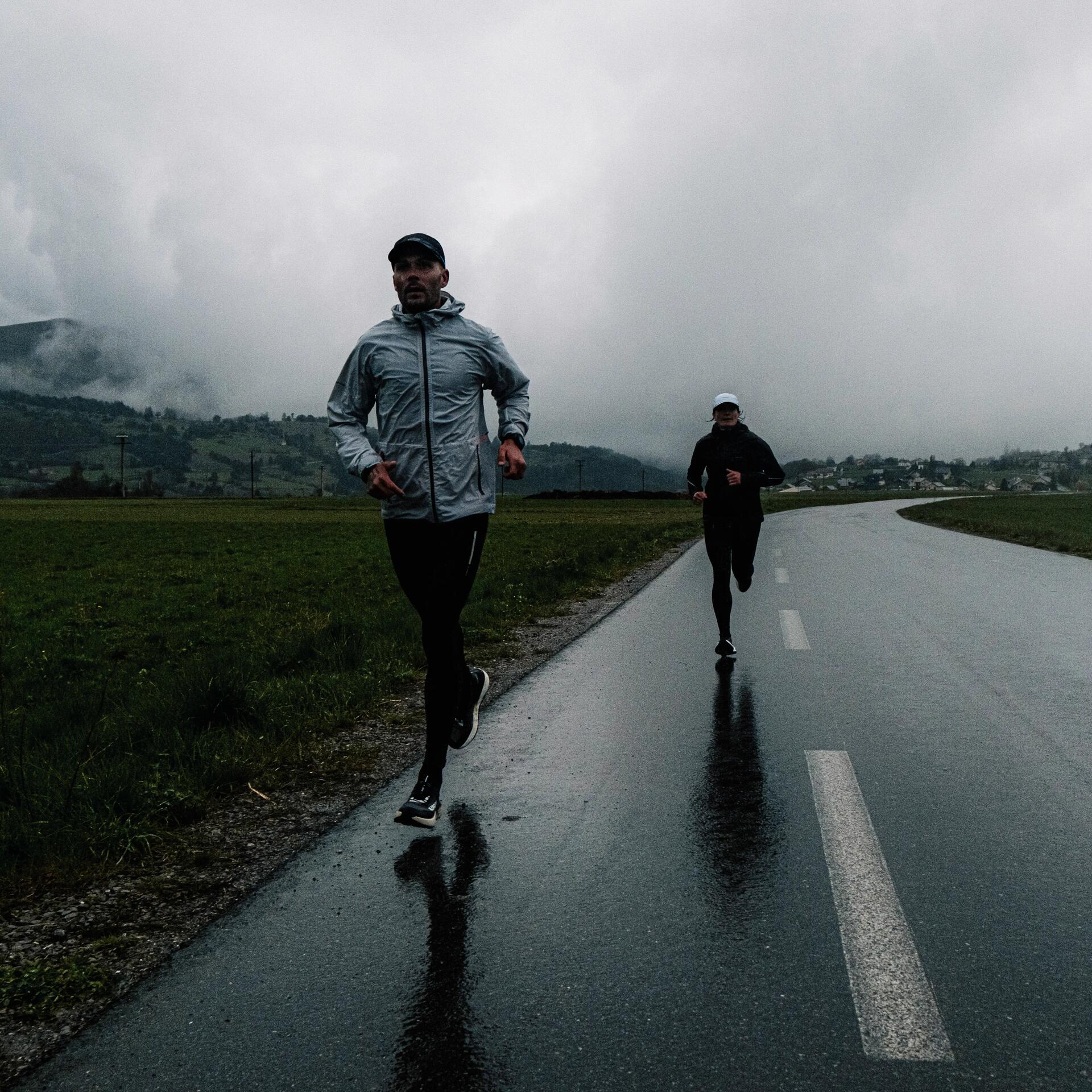 Zwei Jogger laufen auf nasser Asphaltstraße, grauer Himmel, Wolken, grüne Wiese, dunkle Silhouetten spiegeln sich.