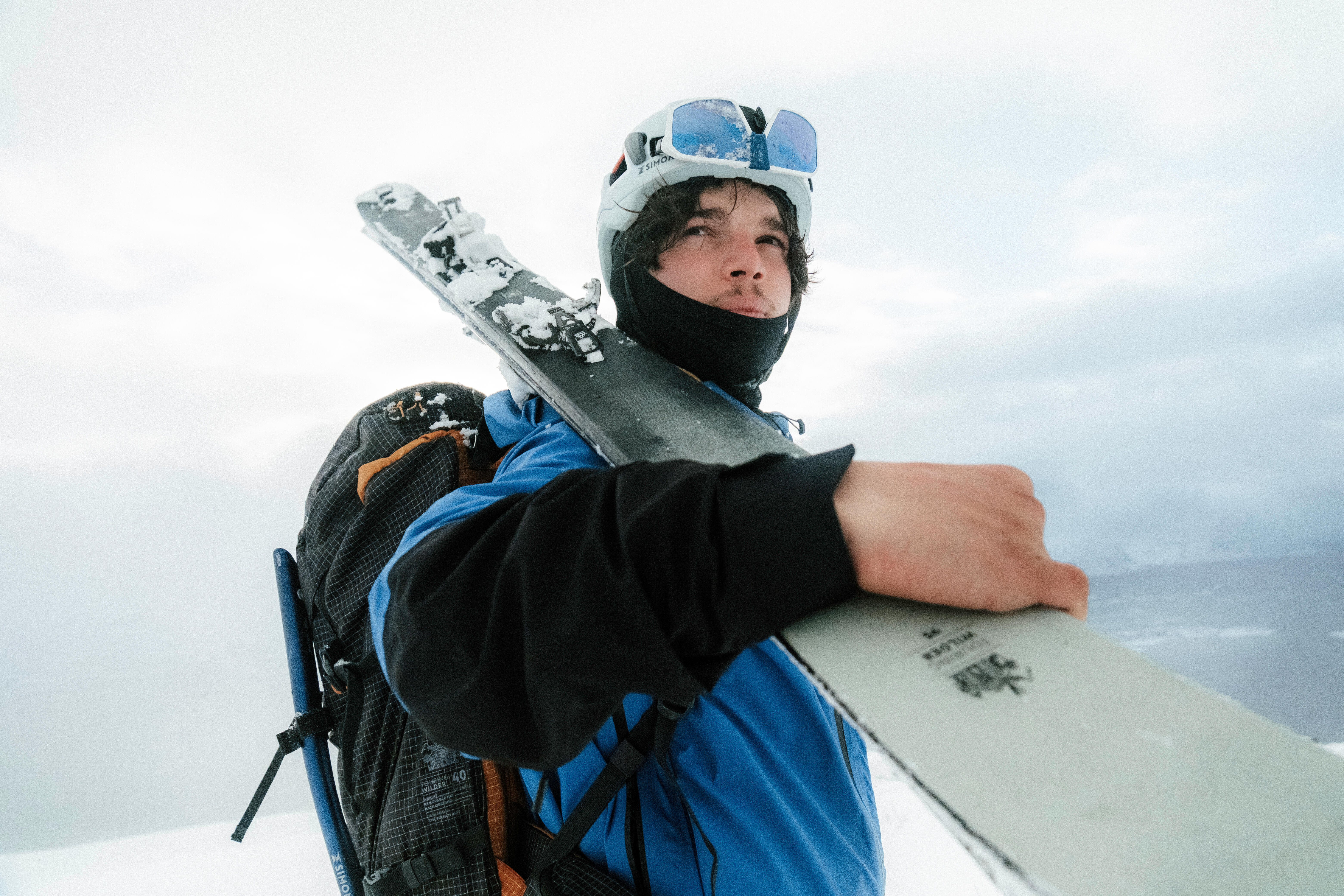 Skier wearing a black ski mask, helmet and goggles while carrying skis