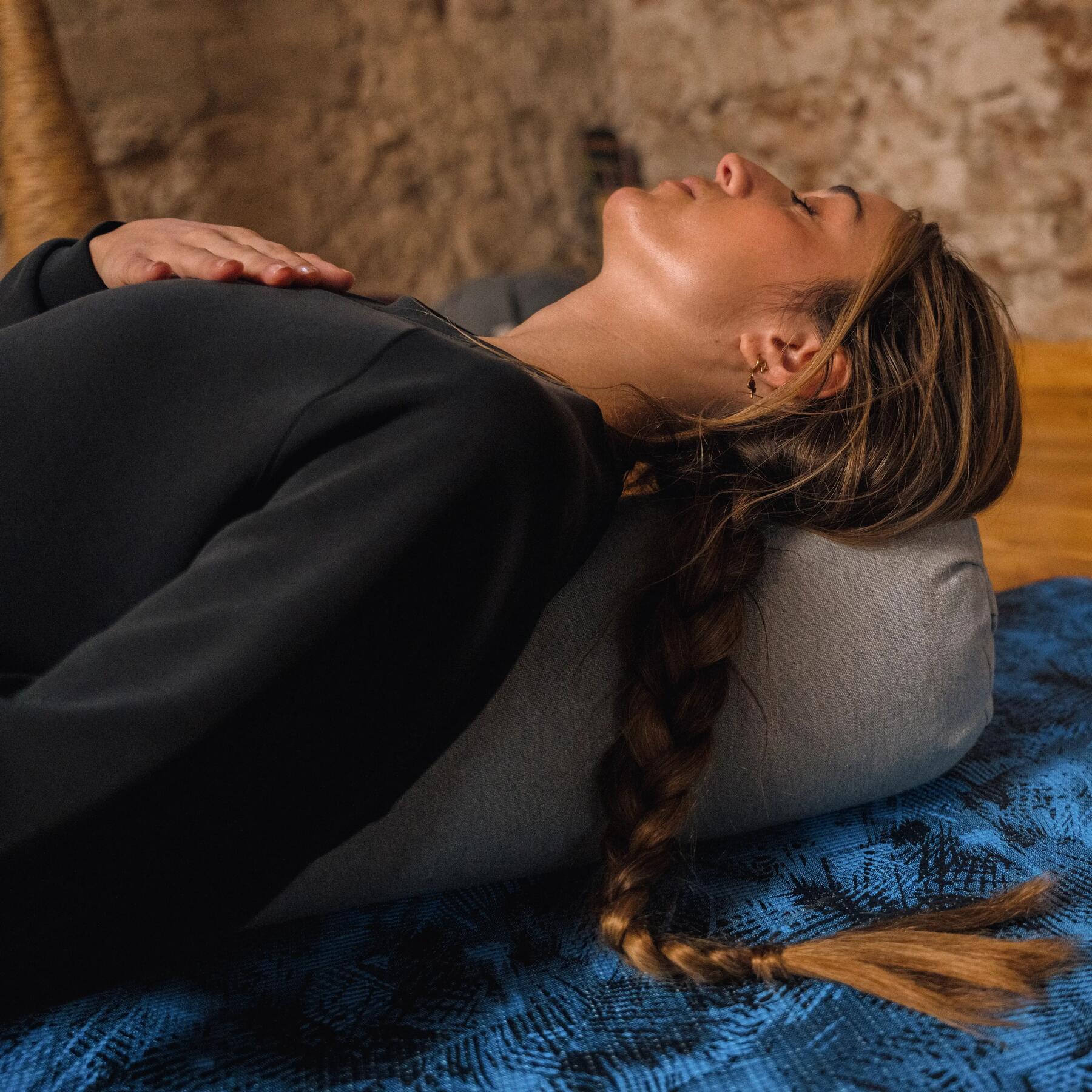 Frau liegt auf dem Boden auf blauer Yogamatte mit grauem Yogakissen unter dem Kopf